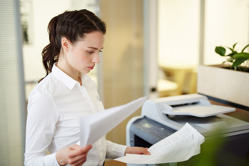 Business woman standing in front of a multifunction printer in office setting looking at documents in hand.