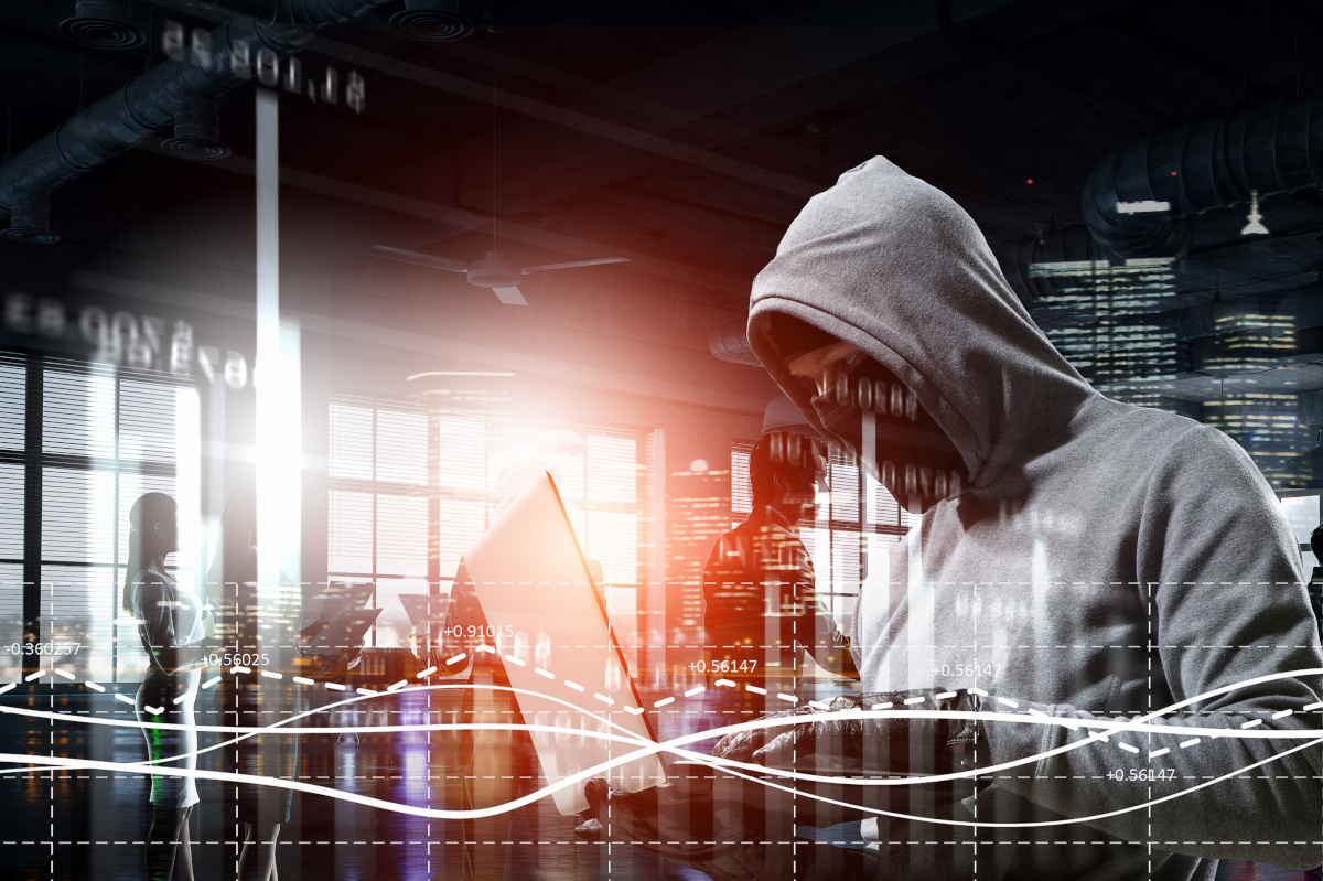 A man in a gray hoodie holding a laptop and typing on it while wearing gloves, with data being displayed over the image to represent a cyber attack